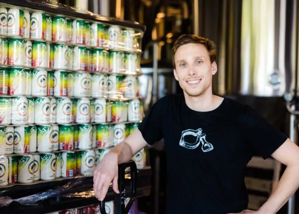 Brewery owner stands with arm resting on cart lift stacked with local brewery cans.