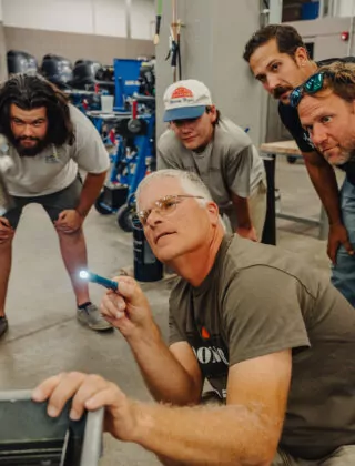 Engineering professor crouched down near car engine and students crowded around listening intently.