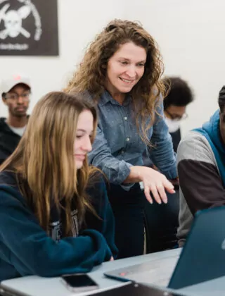 Professor stands behind two students at their desks explaining a class concept.