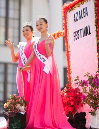 Two women in matching pink gowns stand on a parade float at the Azalea Festival.