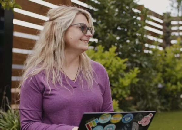 Female banking employee sits in courtyard working on sticker covered laptop.