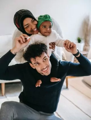 Smiling mother embraces baby sitting on father's shoulders.